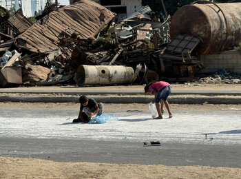 On Salah Ad Din Road in Deir al Balah, Gaza, two children collect flour spilled from a humanitarian truck, barefoot amid the remnants of war, 5 November 2024. Photo by OCHA