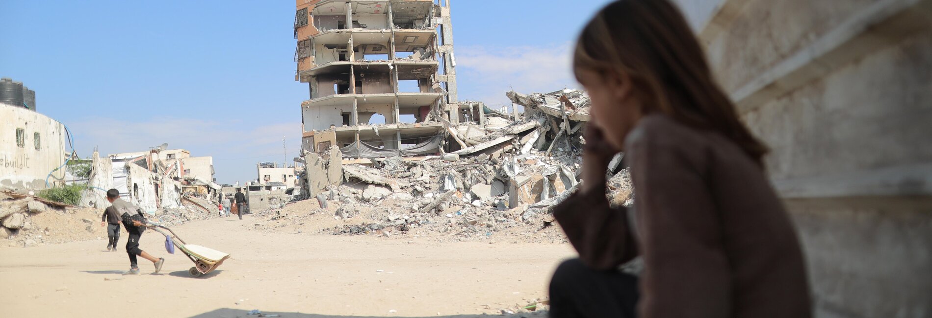 A young girl sitting on the ground with a destroyed building behind her and rubble all around. Photo by UNFPA Palestine/Media Clinic